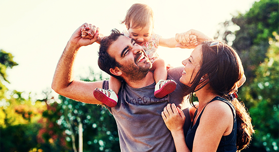 Imagem de uma família, formada por um homem, com um bebê nos ombros e uma mulher. Todos estão felizes e sorrindo. Ao fundo há árvores, como se estivessem em um parque.