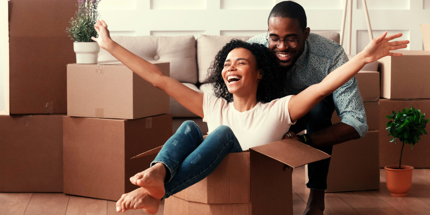 Casal feliz, compradores da sua primeira casa. Ambos estão se divertindo, onde a esposa está sentada em uma caixa de papelão, enquanto o marido a empurra no apartamento novo.