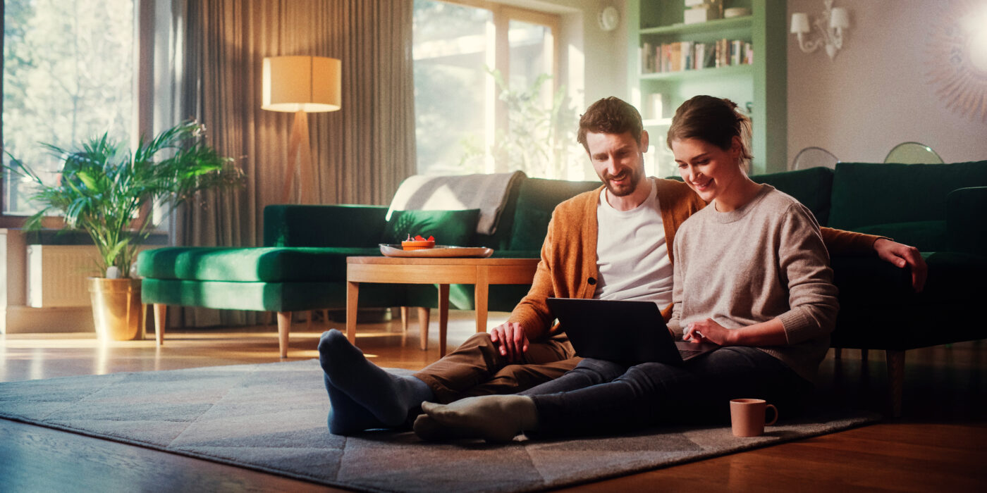 Casal feliz, sentados no chão de uma sala de apartamento, encostados no sofá, utilizando um notebook.